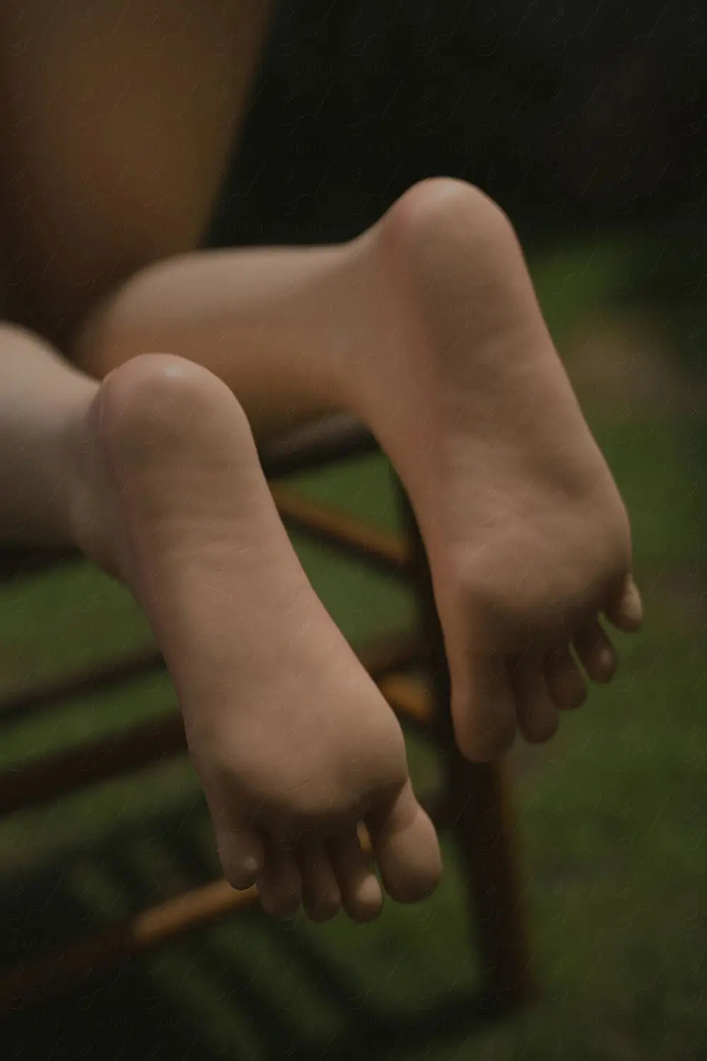 Strider sex doll bare feet close-up, smooth warm-tone TPE skin with natural arch and toe contours, natural unpainted toenails, both feet resting crossed on wooden chair rung with soles facing camera, toes slightly curled, dark moody ambient light with green lawn background – extreme close-up foot detail shot – lifelike realistic sex doll foot detail by TopLoveDoll
