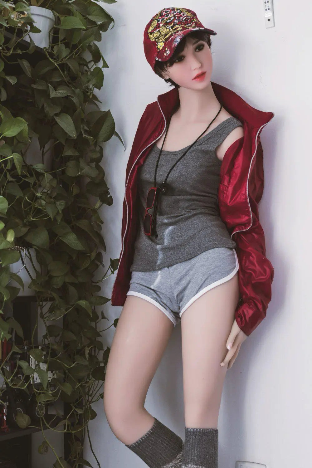 Amira short dark brown hair red lip grey tank denim shorts red jacket tomboy feminine contrast pose, baseball hat, standing next to the plants shelf  – girl-next-door companion sex doll by TopLoveDoll