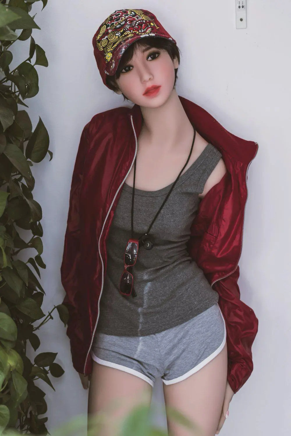 Amira short dark brown hair red lip grey tank denim shorts red jacket tomboy feminine contrast pose, standing next to the plants shelf, baseball hat– girl-next-door companion sex doll by TopLoveDoll
