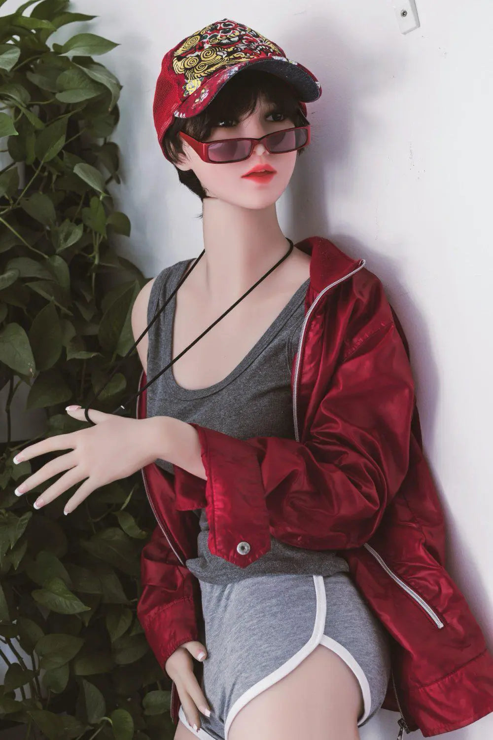 Amira short dark brown hair red lip grey tank denim shorts red jacket tomboy feminine contrast pose, standing against the wall next to the plants shelf, baseball hat, red sunglasses, side view– girl-next-door companion sex doll by TopLoveDoll