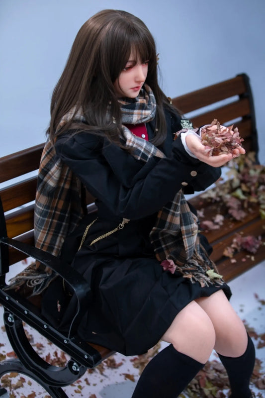 Japaki sex doll serene downcast gaze, dark amber eyes, long straight dark brunette hair with blunt fringe, plaid wool scarf loosely draped, navy blue school blazer with gold buttons, red ribbon bow tie, black pleated mini skirt, black knee-high socks, both hands cupped holding dried autumn leaves, seated on wooden park bench with fallen leaves scattered on ground and pale blue winter sky background – three-quarter full-body seated shot – lifelike Japanese anime girl-next-door sex doll by TopLoveDoll